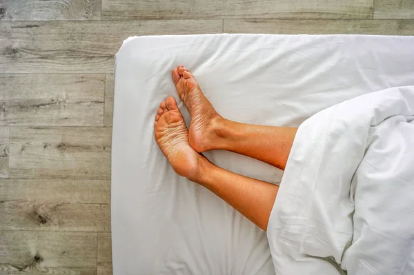 Woman legs on the bed sheet under blanket close-up. Female bare feet on a white sheet, top view, closeup. Closeup naked female legs model onthe bed