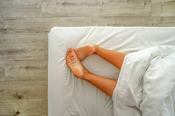 Woman legs on the bed sheet under blanket close-up. Female bare feet on a white sheet, top view, closeup. Closeup naked female legs model onthe bed