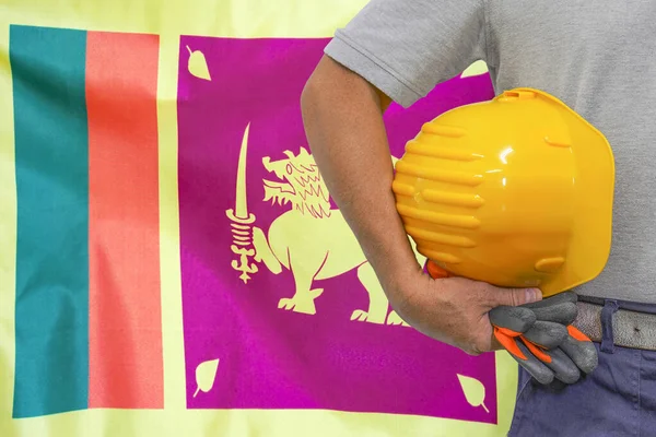 Close-up of hard hat holding by construction worker on Sri Lanka flag background. Hand of worker with yellow hard hat and gloves. Concept of Industry, construction and industrial workers in Sri Lanka