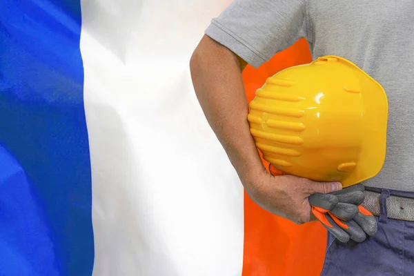 Close-up of hard hat holding by construction worker on France flag background. Hand of worker with yellow hard hat and gloves. Concept of Industry, construction and industrial workers in France