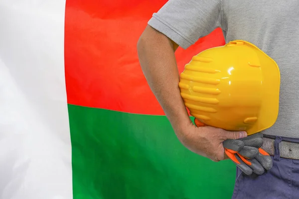Close-up of hard hat holding by construction worker on Madagascar flag background. Hand of worker with yellow hard hat and gloves. Concept Industry, construction and industrial workers in Madagascar