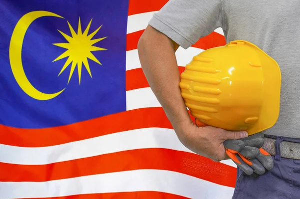 Close-up of hard hat holding by construction worker on Malaysia flag background. Hand of worker with yellow hard hat and gloves. Concept of Industry, construction and industrial workers in Malaysia