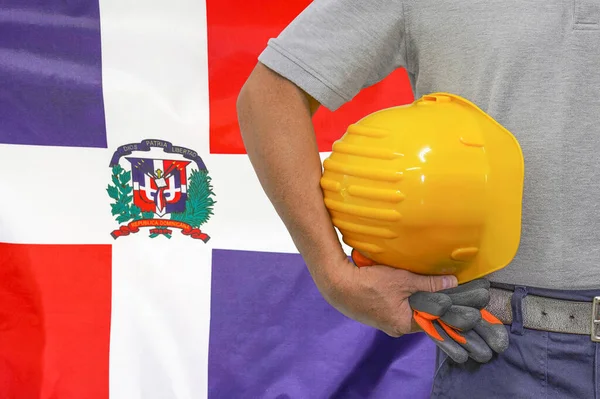 Close-up of hard hat holding by construction worker on Dominican Republic flag background. Hand of worker with yellow hard hat and gloves. Construction and industrial workers in Dominican Republic