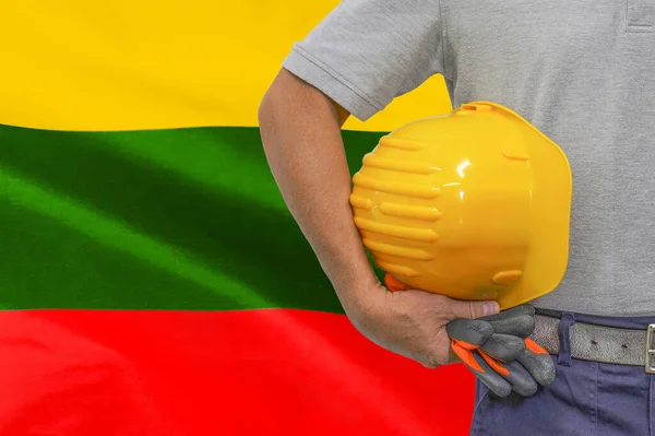Close-up of hard hat holding by construction worker on Lithuania flag background. Hand of worker with yellow hard hat and gloves. Concept of Industry, construction and industrial workers in Lithuania