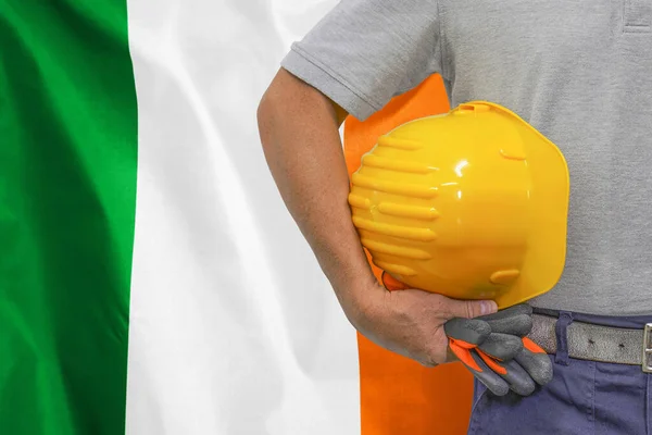 Close-up of hard hat holding by construction worker on Ireland flag background. Hand of worker with yellow hard hat and gloves. Concept of Industry, construction and industrial workers in Ireland