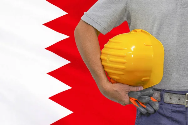 Close-up of hard hat holding by construction worker on Bahrain flag background. Hand of worker with yellow hard hat and gloves. Concept of Industry, construction and industrial workers in Bahrain