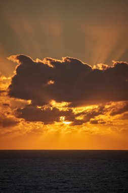 Bulutların arkasındaki güzel güneş sabahın erken saatlerinde Atlantik Okyanusu 'nun üzerinde ışıl ışıl yükselir ve gökyüzünü ve bulutları parlak turuncu renkler ve ışıklarla boyar.