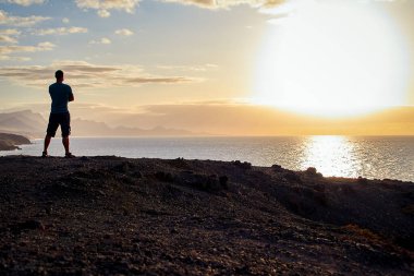 Man in a blue t-shirt enjoys the view of the sea in the evening light and the sunset on the coast of Fuerteventura at the golden hour