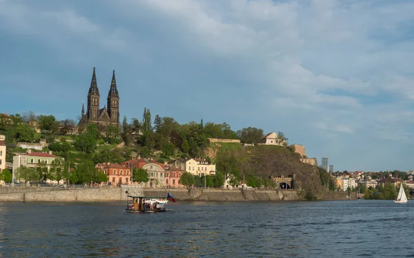 小さなフェリーボートと歴史的な砦を持つヴルタヴァ川の銀行ヴィシェフラッド聖ピーターとポール大聖堂、緑の丘と岩の上にゴシック様式の大聖堂教会、プラハ、チェコ共和国、春の日. — ストック写真