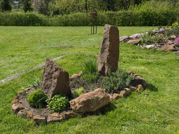 Vista sobre jardín de primavera con cama de flores ovalada con hierbas verdes tomillo, lavanda, menta, cebollino y bálsamo dulce con gran roca arenisca y piedras sobre el fondo de hierba verde exuberante —  Fotos de Stock