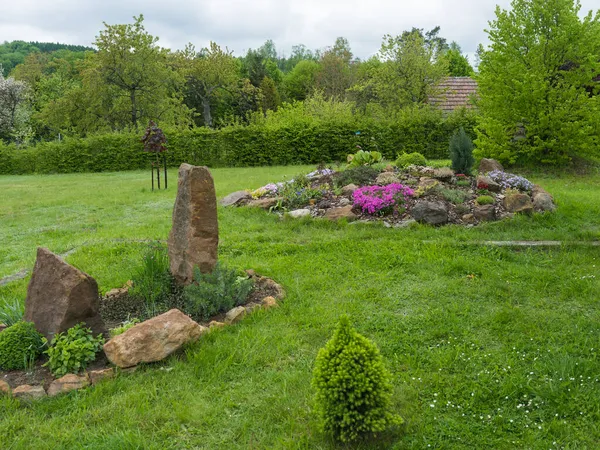 Uitzicht op de lentetuin met ovaal bloembed met groene kruiden met grote zandsteen rots en bloeiende rotstuin op de achtergrond van weelderig groen gras — Stockfoto