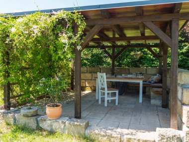 Sıcak kahverengi kaplan ahşap çardak, beyaz masalı pergola, yastıklı sandalyeler ve kumtaşı duvarlı battaniye ve tırmanma bitkileri ve çiçek açan pembe çömlek zengini. Güneşli yaz günü