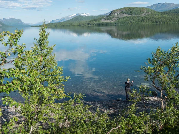 Pescador no rio Lule, em claro calmo lago barragem Lulealven água doce em Saltoluokta, na Suécia Lapônia no dia ensolarado de verão. Montanha rochosa verde, bétulas e nuvens. — Fotografia de Stock