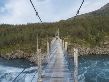 Vuojatadno nehri üzerindeki tahta asma köprü Laponya İsveç arktik ormanının arka planında, yaz doğal manzarasında akıntıya kapılır. Dar köprü mesafeye gidiyor. Perspektif görünümü.