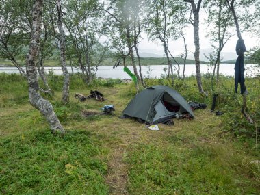 Small open green tent with backpack and hiking gear on grassy shore of Tarra river with birch trees. Swedish Lapland Landscape with green hills at Padjelantaleden hiking trail. Summer day