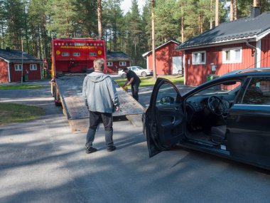Jokkmokk, Norrbotten, Sweden, Agust 17, 2021: Two men tow truck drivers loading black car onto a tow truck at Arctic Camp Jokkmokk with red wooden cabins.