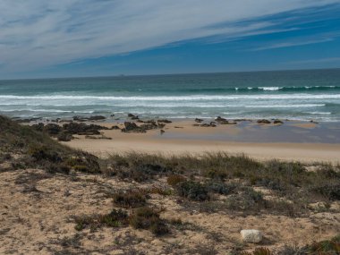 Porto Covo, Portekiz yakınlarındaki vahşi Rota Vicentina sahilinde okyanus dalgaları, keskin kayalar, taşlar ve yeşil bitki örtüsü olan boş küçük kumsal manzarası