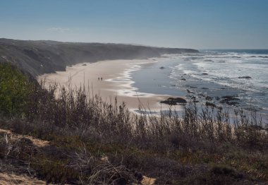 Praia do Brejo Largo sahilinin hava manzarasında iki uzak yürüyüşçü figürü, okyanus dalgaları ve keskin taşlar, kayalıklar ve Rota Vicentina sahilinde kum. Vila Nova de Milfontes, Portekiz. Güneşli gün, mavi gökyüzü