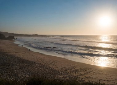 Praia Grande de Almograve kumsalında uzak siluet insanlar ve okyanus dalgaları altın saat ışığı, açık mavi gökyüzü. Rota Vicentina Sahili, Almograve, Portekiz.