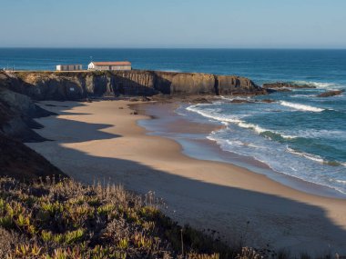 Praia Grande de de Almograve kumsal manzarası. Okyanus dalgaları, uçurum, yeşil bitki örtüsü ve küçük sahil evi. Açık mavi gökyüzü. Rota Vicentina Sahili, Almograve, Portekiz.