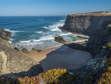 Portekiz, Almograve yakınlarındaki Rota Vicentina sahilindeki boş Praia do Cavaleiro plajı sarp kayalıkları, okyanus dalgaları, taşlar, ıslak altın kum ve yeşil bitki örtüsü manzarası