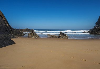 Portekiz 'in Almograve yakınlarındaki vahşi Rota Vicentina sahilinde sarp kayalıkları, okyanus dalgaları, taşları ve ıslak altın kumu olan boş Praia do Cavaleiro plajı manzarası. Açık mavi gökyüzü, boşluğu kopyala.