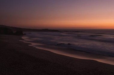 Kum sahili Praia Grande de Almograve 'in pembe turuncu ve mor ışıkta bulanık okyanus dalgalarıyla esrarengiz uzun görüntüsü. Günbatımından sonraki mavi saat Rota Vicentina Sahili, Almograve, Portekiz.