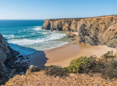Okyanus dalgaları, uçurumlar ve taşlarla dolu boş Praia do Tonel plajı, Portekiz, Zambujeira yakınlarındaki vahşi Rota Vicentina sahilinde ıslak altın kum ve yeşil bitki örtüsü