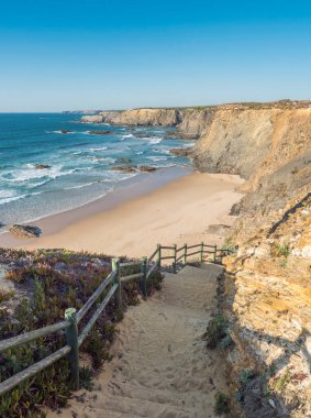 Okyanus dalgaları, uçurumlar ve taşlarla dolu boş Praia de Nossa Senhora plajı, Portekiz 'in Zambujeira do Mar yakınlarındaki vahşi Rota Vicentina sahilinde ıslak altın kum ve yeşil bitki örtüsü.
