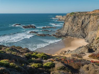 Boş Praia da Baia da Arquinha plajı okyanus dalgaları, uçurumlar ve taşlar, ıslak altın kum ve yeşil bitki örtüsü Zambujeira yakınlarındaki Rota Vicentina sahilleri, Mar, Portekiz