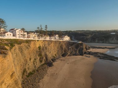 Zambujeira do Mar 'ın kumsaldan görünen beyaz evleri Praia da Zambujeira do Mar, kayalıkları ve kayaları olan deniz kıyıları, güneşli bir gün, açık mavi gökyüzü. Rota Vicentina Sahili, Portekiz.