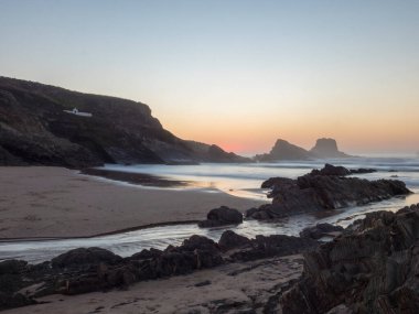 Gün batımından sonra Praia da Zambujeira do Mar in rock and cliff, little stream ve bulanık okyanus dalgaları pembe ışıkta. Rota Vicentina Sahili, Odemira, Portekiz
