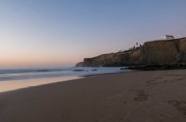 Gün batımından sonra Praia da Zambujeira 'nın küçük şapeli ve pembe ışıkta bulanık okyanus dalgalarıyla Mar' ı yaptığı kumsalın mavi saati. Rota Vicentina Sahili, Odemira, Portekiz.