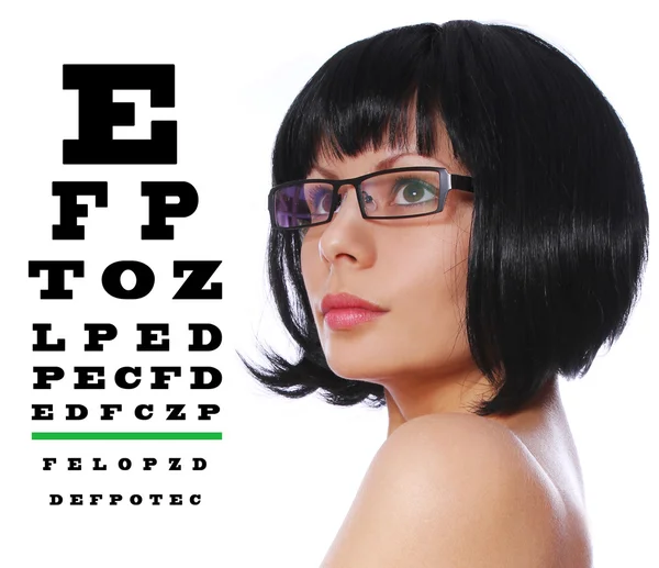 Opticida. Linda morena vestindo óculos e Snellen olho exame gráfico isolado no fundo branco — Fotografia de Stock