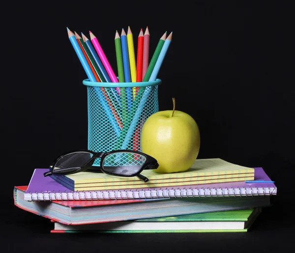 Torniamo al concetto di scuola. Una mela, matite colorate e bicchieri su mucchio di libri su sfondo nero . — Foto Stock