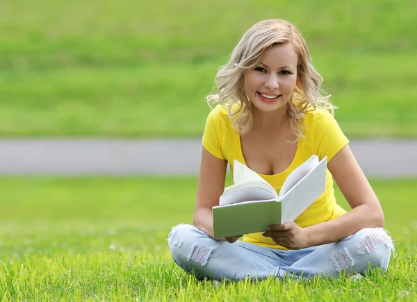 Mädchen lesen das Buch. glückliche blonde schöne junge Frau mit Buch auf dem Gras sitzend. Draußen. Blick in die Kamera — Stockfoto