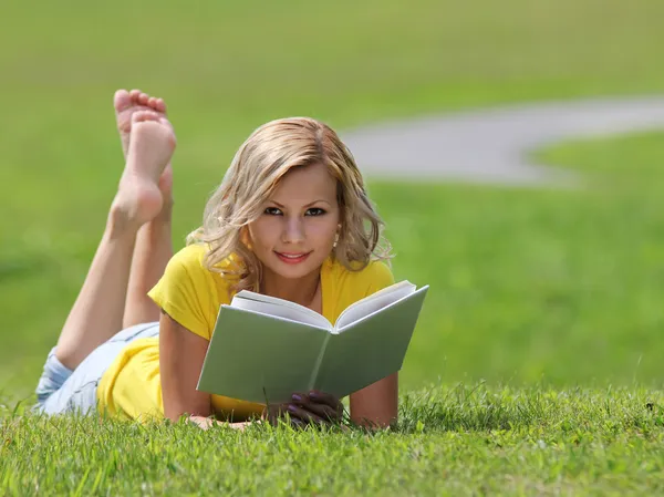 Девушка читает книгу. Блондинка красивая молодая женщина с книгой, лежащей на траве. На улице. Солнечный день. Возвращайся в школу. Смотреть в камеру — стоковое фото