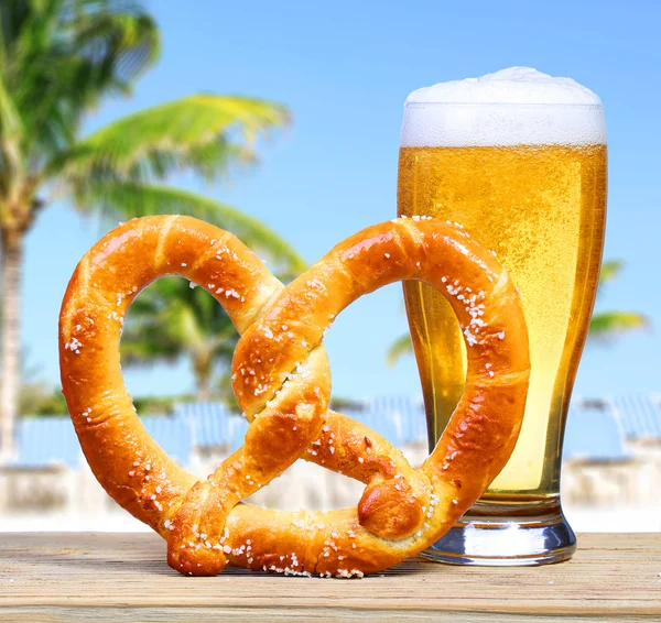 Copo de cerveja com Pretzel alemão sobre vista para a praia com palmas — Fotografia de Stock