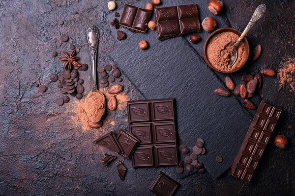 Pieces of broken tasty milk and dark chocolate bars with cocoa beans and hazelnut on black slate board and concrete background