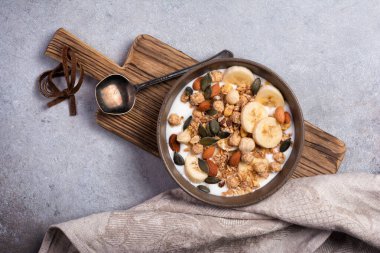 Yoğurtlu granola, taze muz, badem ve fındık, sağlıklı kahvaltı için balkabağı tohumları gri beton arka planda.