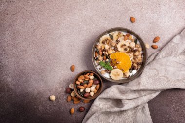 Yoğurtlu granola, taze muz, portakal, badem ve fındık, kahverengi beton zemin üzerinde sağlıklı kahvaltı için kabak çekirdeği.