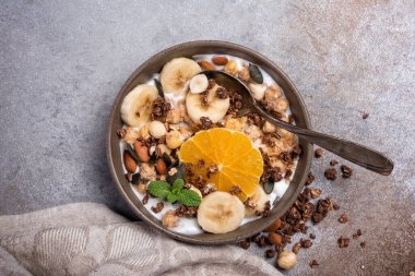 Yoğurtlu granola, taze muz, portakal, badem ve fındık, sağlıklı kahvaltı için balkabağı tohumları gri beton arka planda.