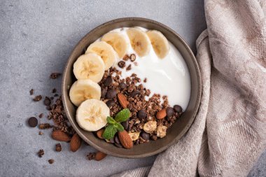 Yoğurtlu granola, taze muz, çikolata damlaları ve badem fıstıklı sağlıklı kahvaltı için gri beton arka planda