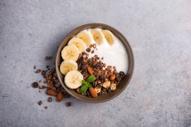 Yoğurtlu granola, taze muz, çikolata damlaları ve badem fıstıklı sağlıklı kahvaltı için gri beton arka planda