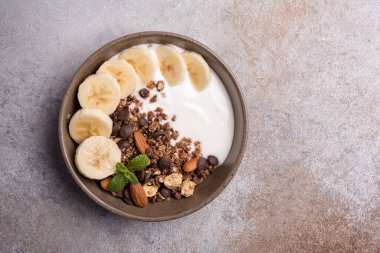 Yoğurtlu granola, taze muz, çikolata damlaları ve badem fıstıklı sağlıklı kahvaltı için gri beton arka planda