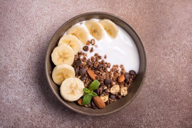 Yoğurtlu granola, taze muz, çikolata damlaları ve badem fıstıklı sağlıklı kahvaltı için kahverengi beton arka planda