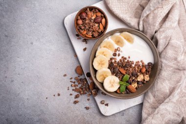 Yoğurtlu granola, taze muz, çikolata damlaları ve badem fıstıklı sağlıklı kahvaltı için gri beton arka planda