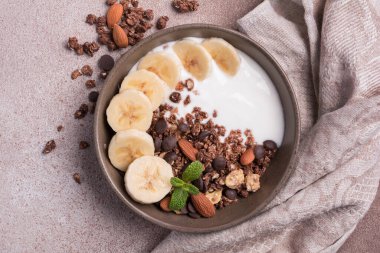 Yoğurtlu granola, taze muz, çikolata damlaları ve badem fıstıklı sağlıklı kahvaltı için kahverengi beton arka planda