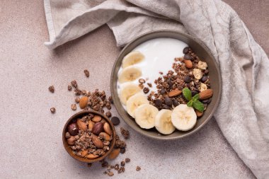 Yoğurtlu granola, taze muz, çikolata damlaları ve badem fıstıklı sağlıklı kahvaltı için kahverengi beton arka planda