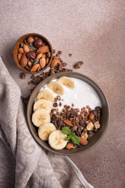 Yoğurtlu granola, taze muz, çikolata damlaları ve badem fıstıklı sağlıklı kahvaltı için kahverengi beton arka planda